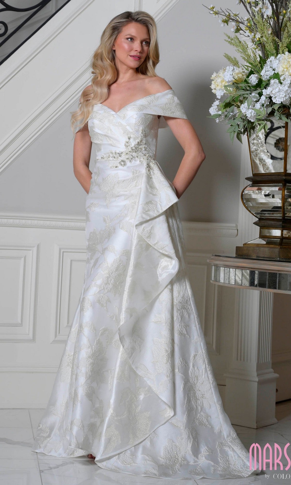 An elegantly dressed woman stands indoors in the ivory Marsoni MV1401 long formal dress with a fitted bodice and draped overlay, near a table topped with a decorative flower arrangement.